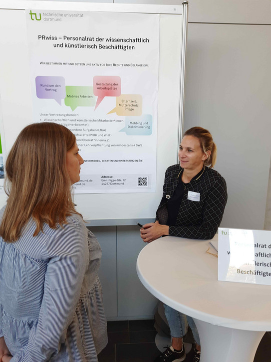 Dr. Stephanie Steden informiert eine Besucherin des Markts der Informationen über die Arbeit des Personalrats der wissenschaftlich und künstlerisch Beschäftigten der TU Dortmund. Im Hintergrund ein Poster mit den wichtigsten Arbeitsfeldern des PRwiss. 