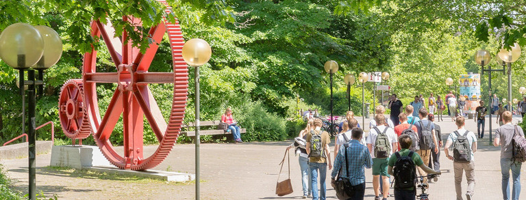 Studierende am Campus Nord Studierende laufen über den Campus Nord. Es ist eine große rote Skulptur in Form von großen Zahnrädern zu erkennen.